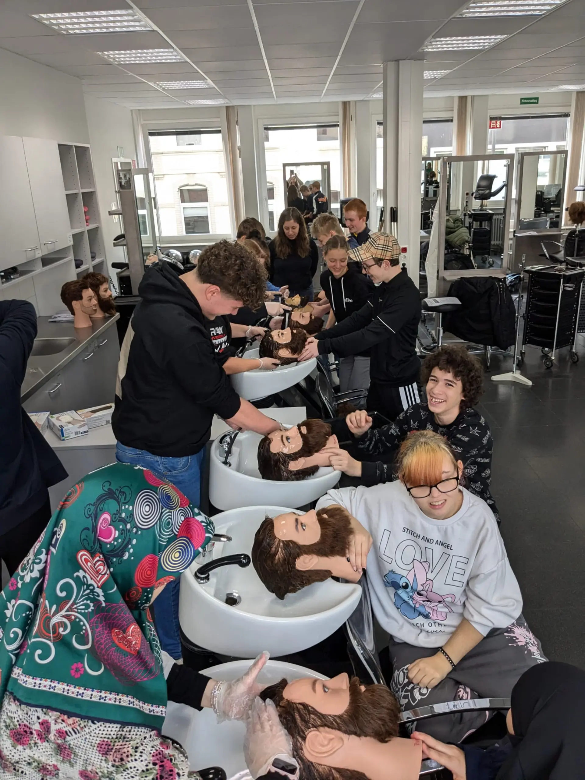 Eine große Gruppe von Schüler*innen probieren in einem Friseursalon aus, wie man die Haare einer fremden Person am Friseursalon-Waschbecken wäscht. Sie üben an Plastikköpfen mit kurzen Haaren. 
