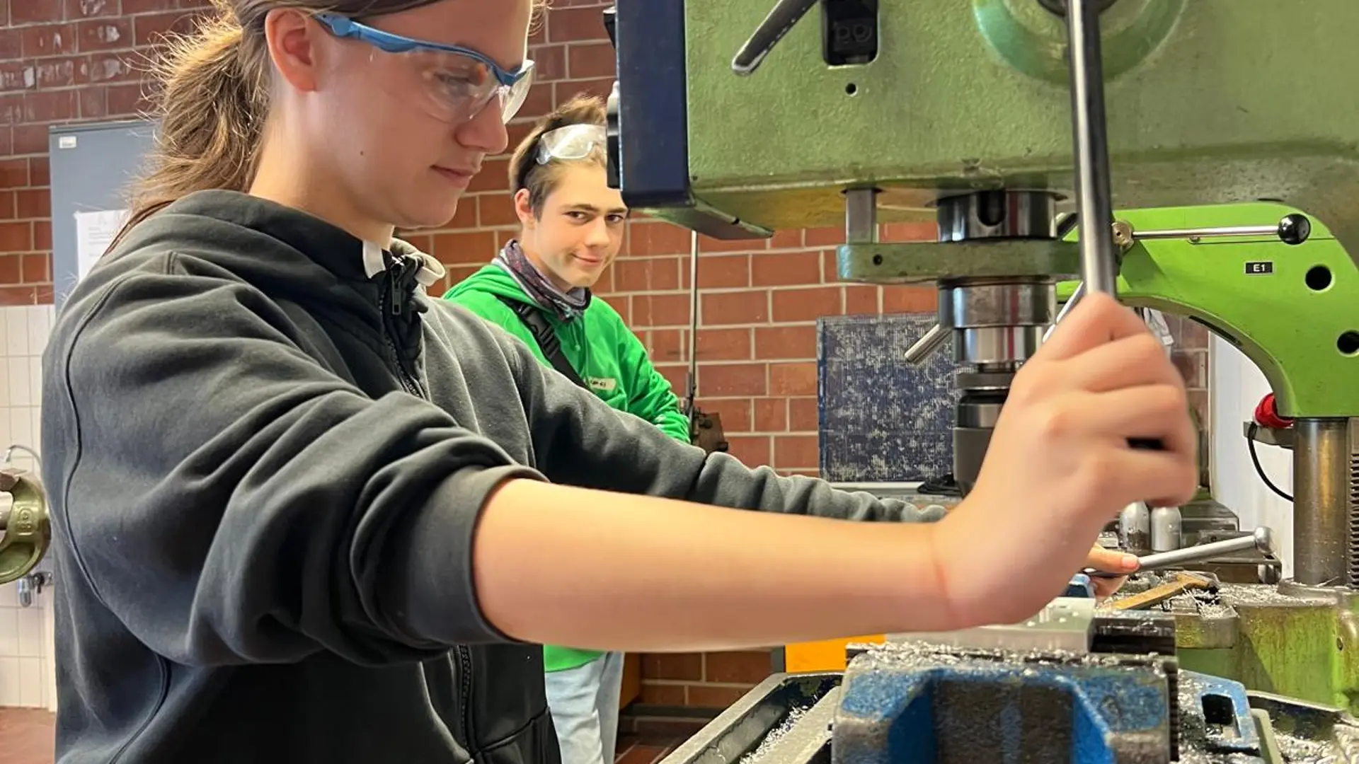 Ein Schüler bedient konzentriert eine Standbohrmaschine in einer Holzwerkstatt. Dabei trägt er eine Schutzbrille.