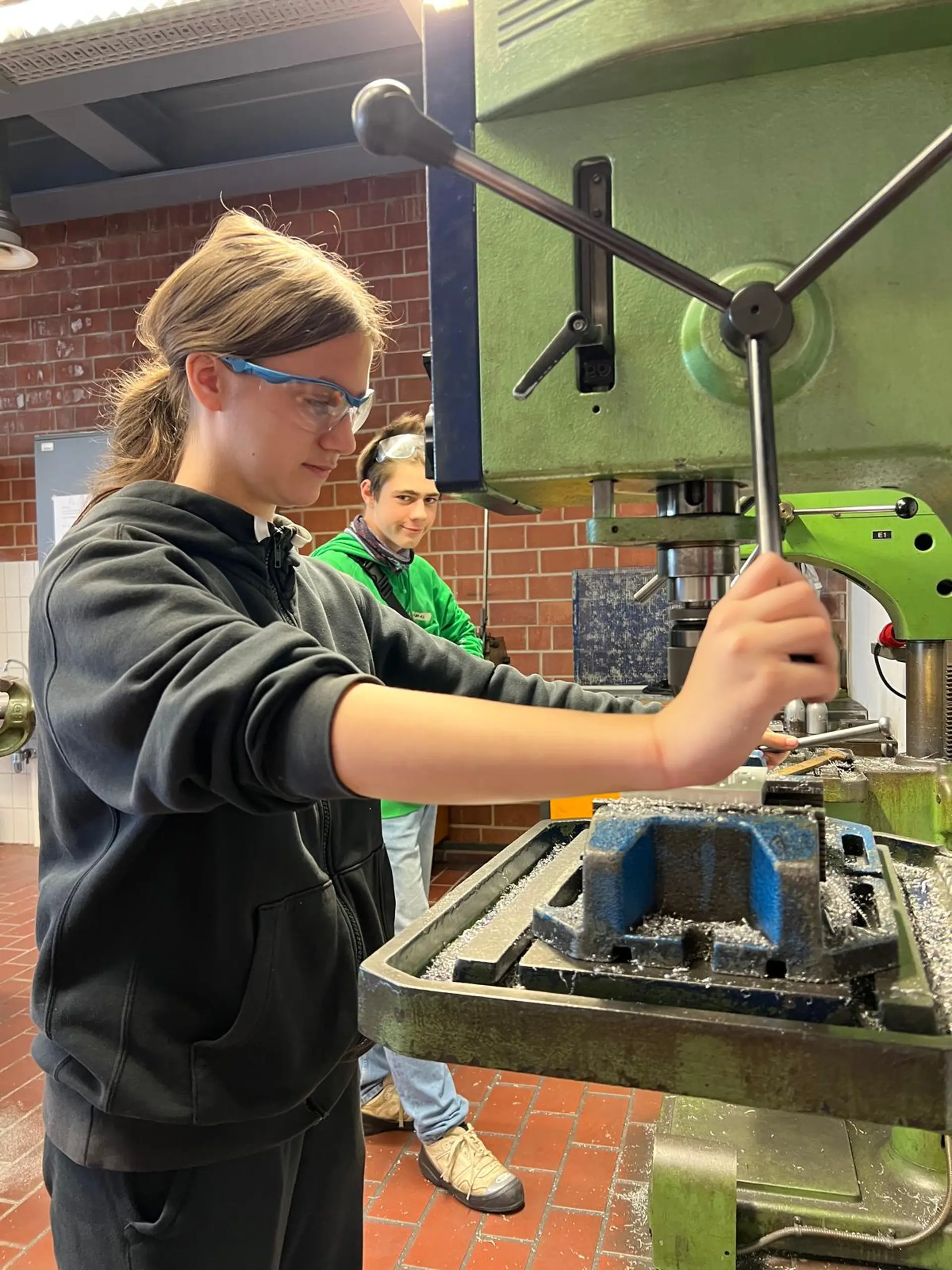 Ein Schüler bedient konzentriert eine Standbohrmaschine in einer Holzwerkstatt. Dabei trägt er eine Schutzbrille.