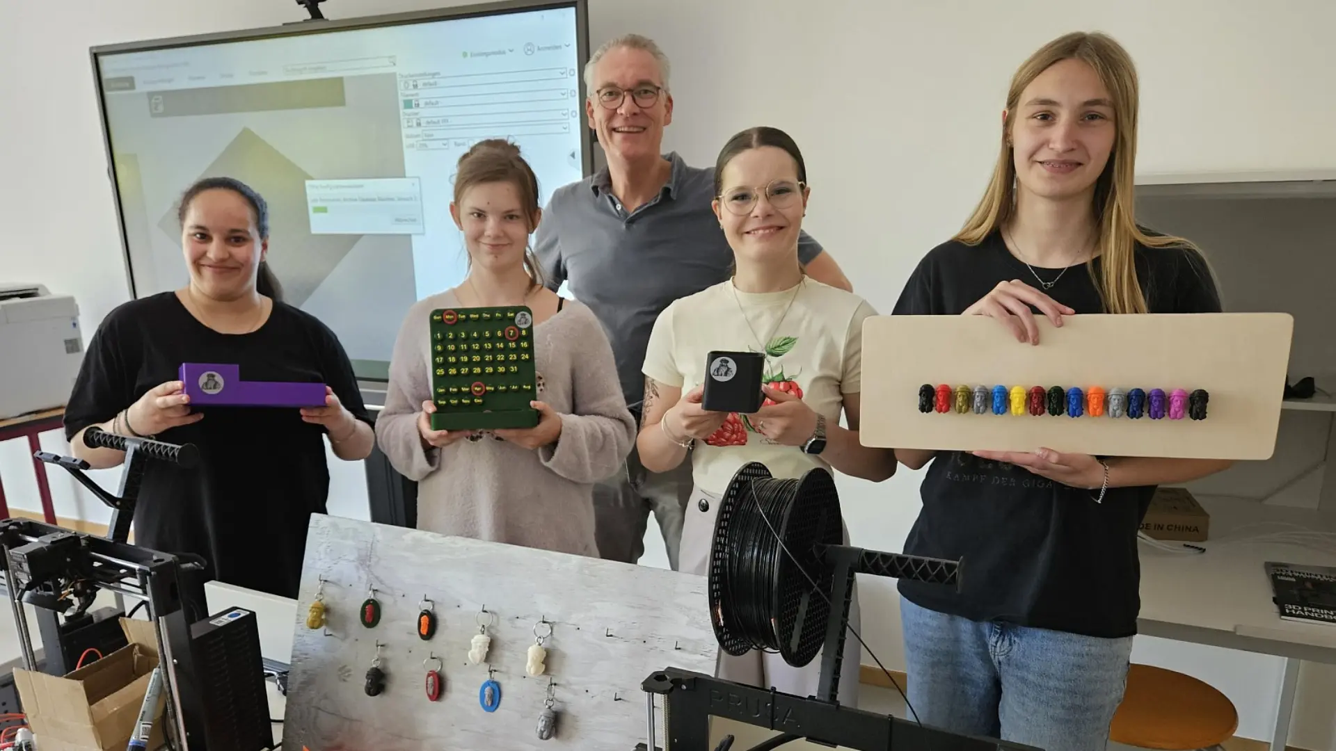 Drei Schülerinnen und eine Lehrkraft stehen im Klassenzimmer und präsentieren ihre 3D-Projekte. Vor ihnen auf den Tischen sind 3D-Drucker, hinter ihnen eine eingeschaltete digitale Tafel zu sehen. 