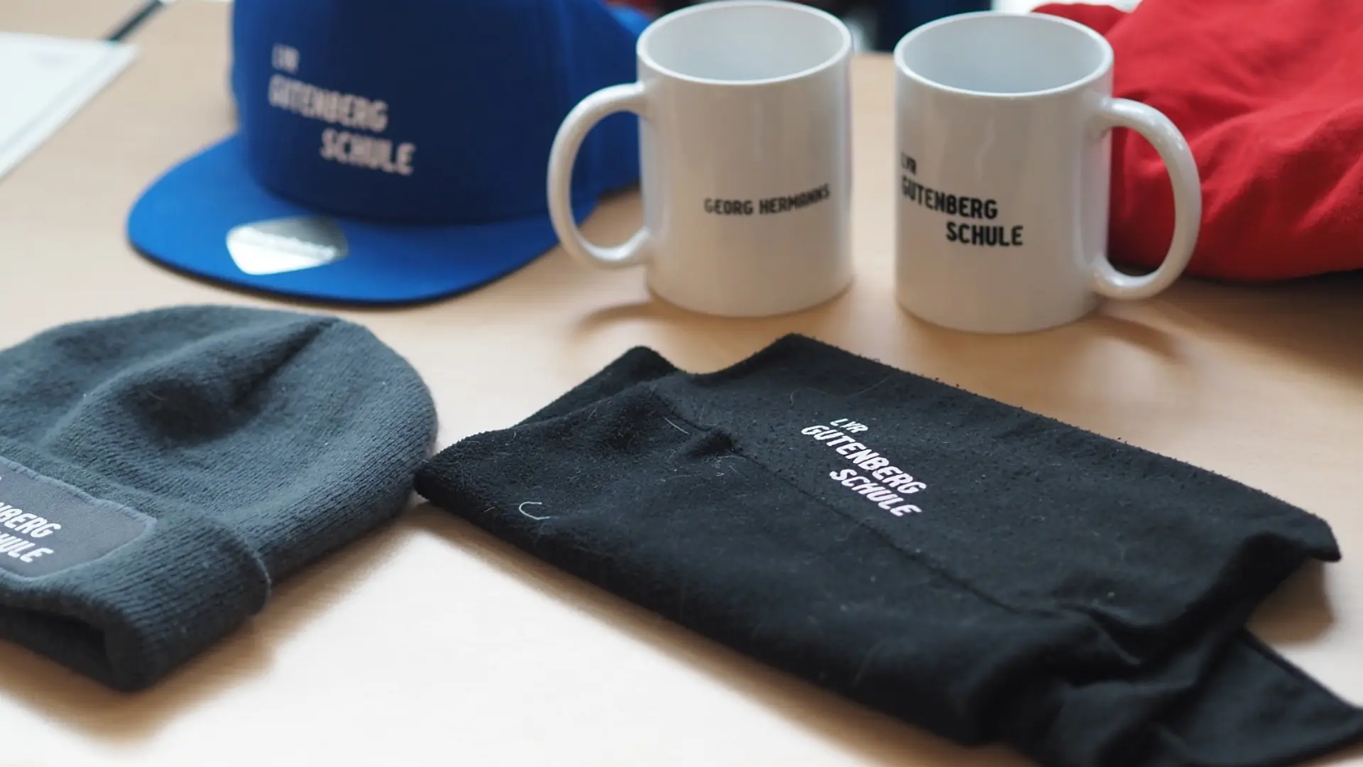 Verschiedene Werbeprodukte wie T-Shirt, Tassen und Caps mit dem ausgeschriebenen Namen der Schule liegen auf einer Tischplatte.