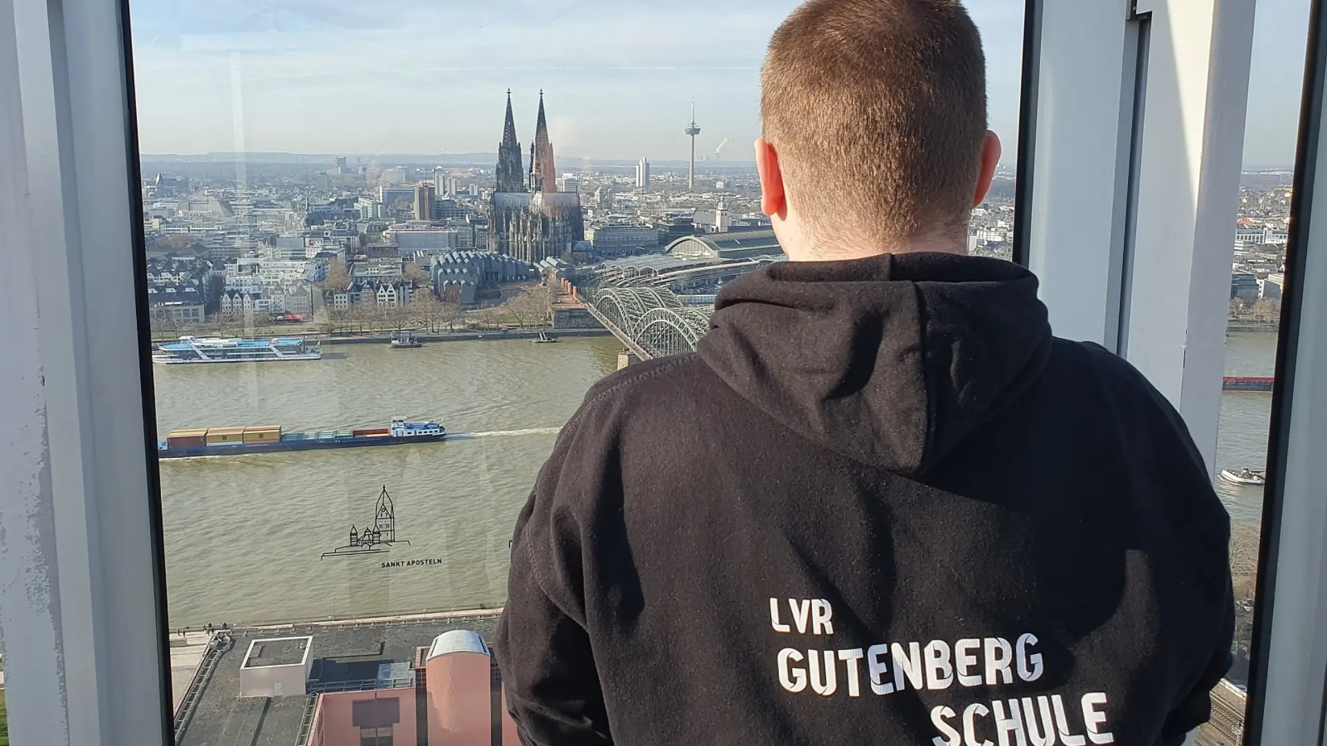 Ein Jugendlicher steht oben auf der Aussichtsplattform im Kölner Triangle Turm und schaut durch das Sicherheitsglas zum Rhein und dem Dom auf der anderen Flussseite.