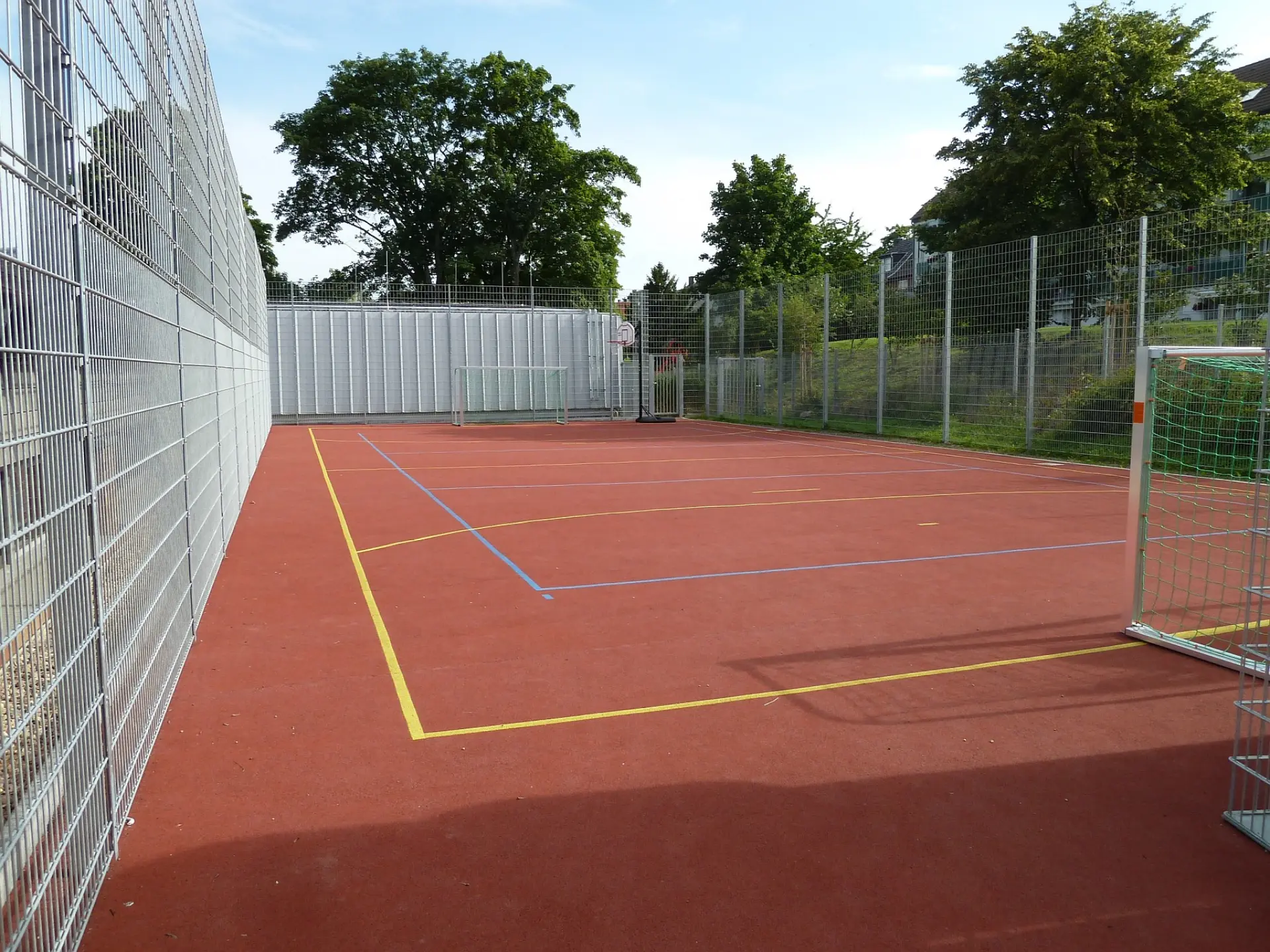 Eingezäunter Allwetter-Sportplatz mit rotem Bodenbelag, Fußballtoren und einem Basketballkorb unter freiem Himmel