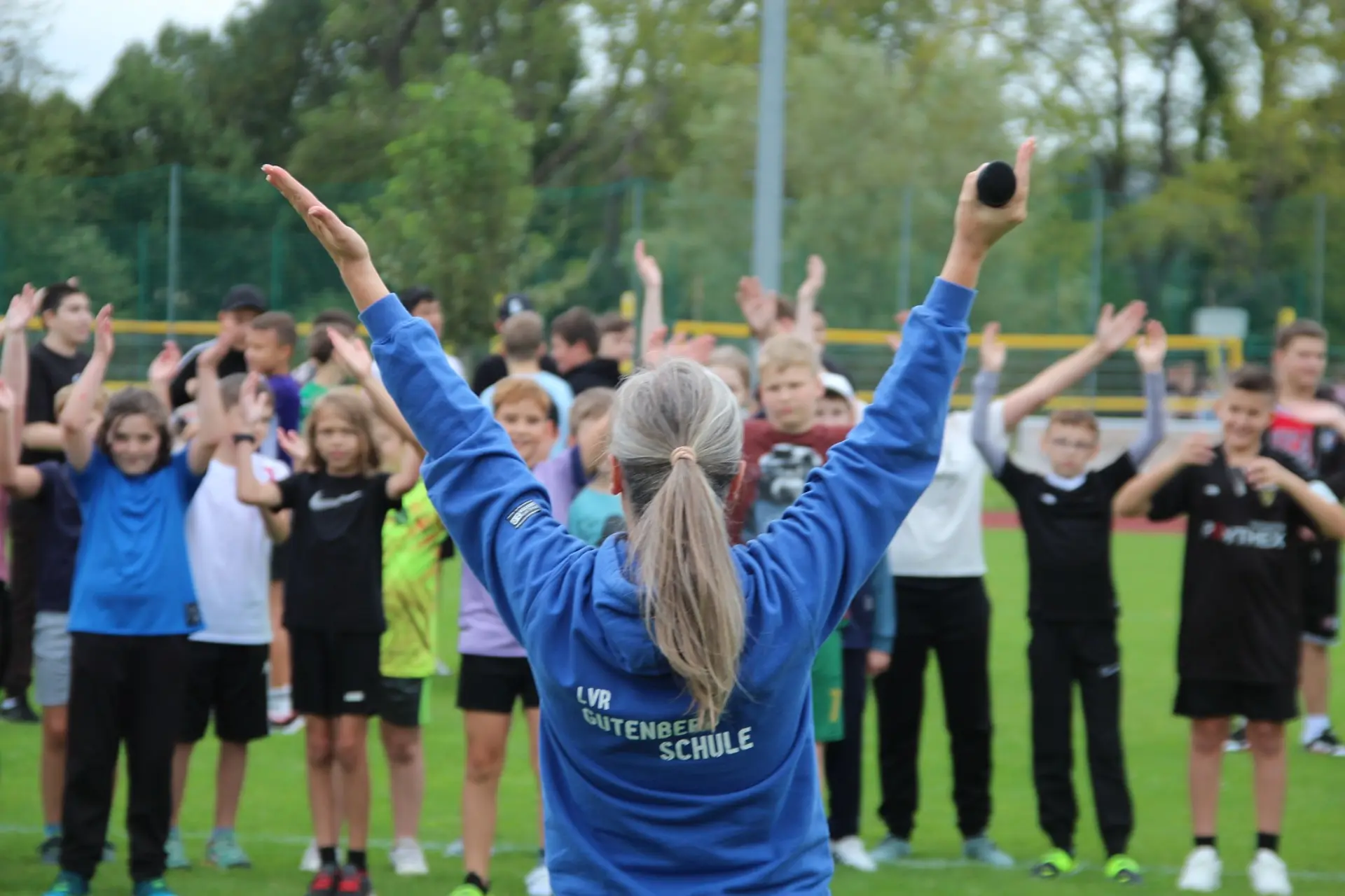 Eine Lehrerin wird von hinten aufgenommen, während sie Aufwärmübungen mit einer Klasse auf dem Sportrasen macht.