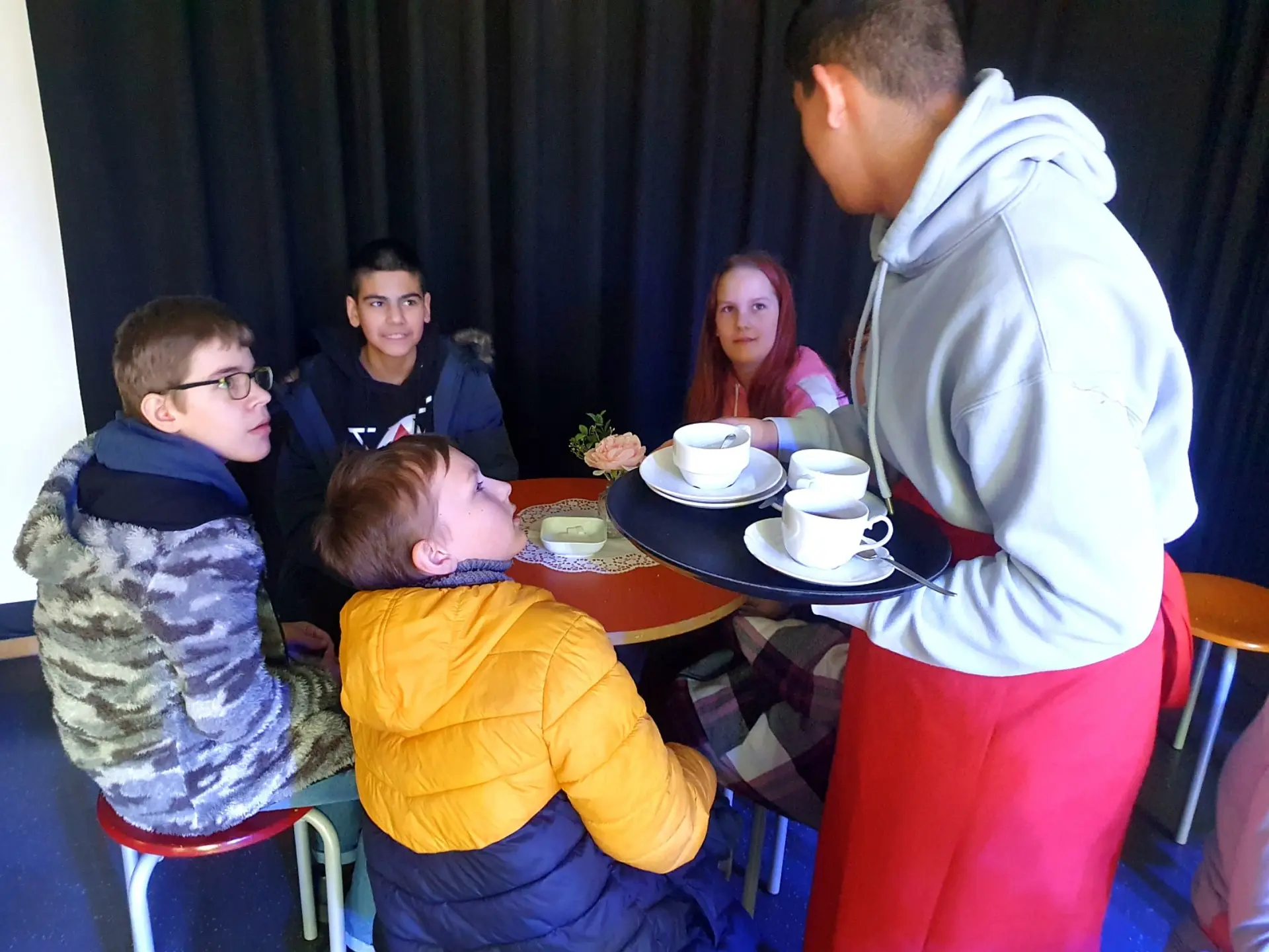Ein Schüler trägt ein Tablett mit drei leeren Kaffeetassen darauf. Er spricht mit vier Schüler*innen, die an den runden Tischen in der Cafeteria sitzen und gerade ihren Tee zu Ende getrunken haben.