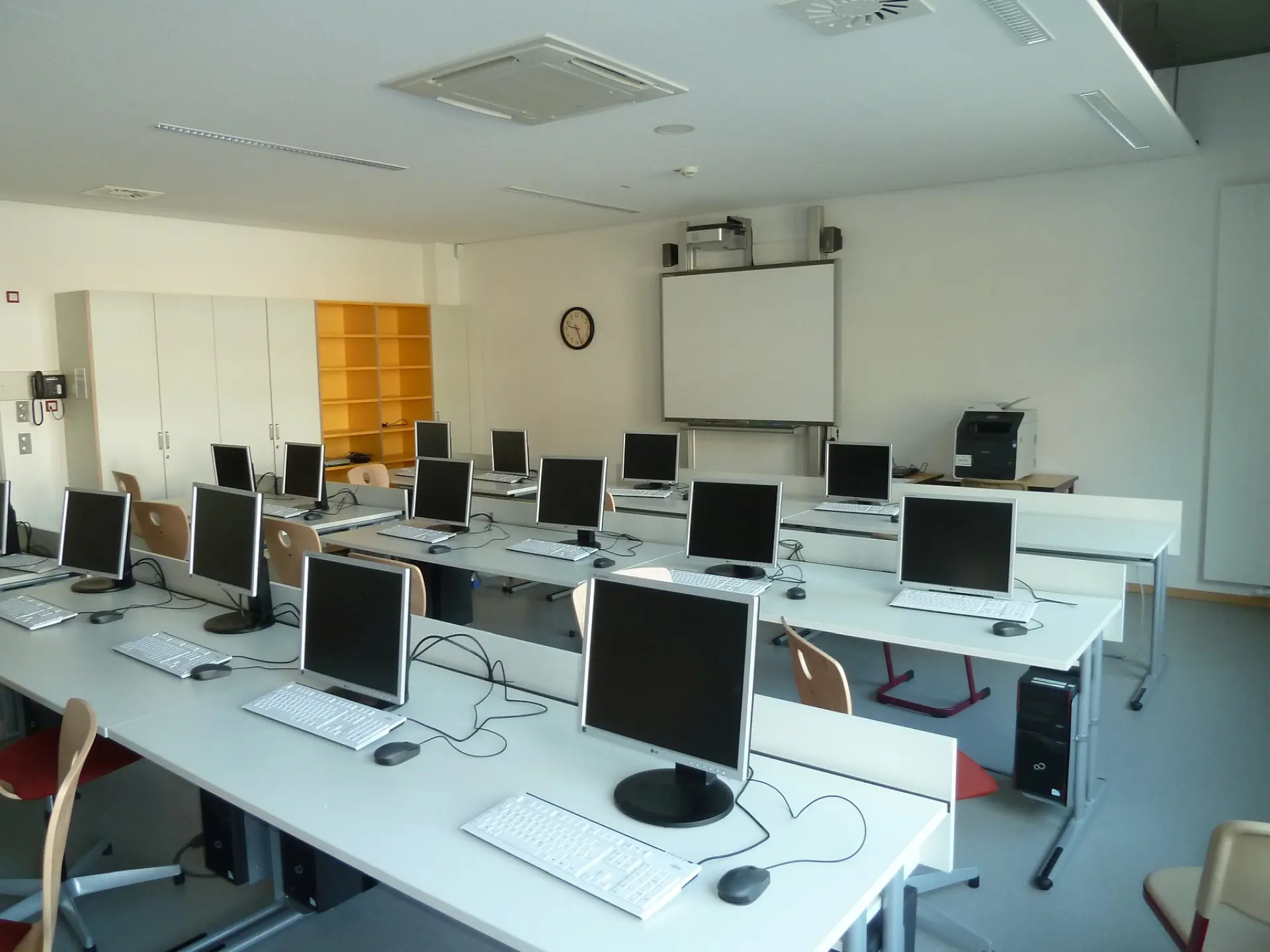 Blick in einen Computerraum der Schule: Mehrere Reihen weißer Arbeitstische mit Desktop-PCs, Monitoren und Tastaturen sind im Raum angeordnet. An der Stirnseite befinden sich ein interaktives Whiteboard, ein Laserdrucker sowie helle Aufbewahrungsschränke mit gelben Akzenten