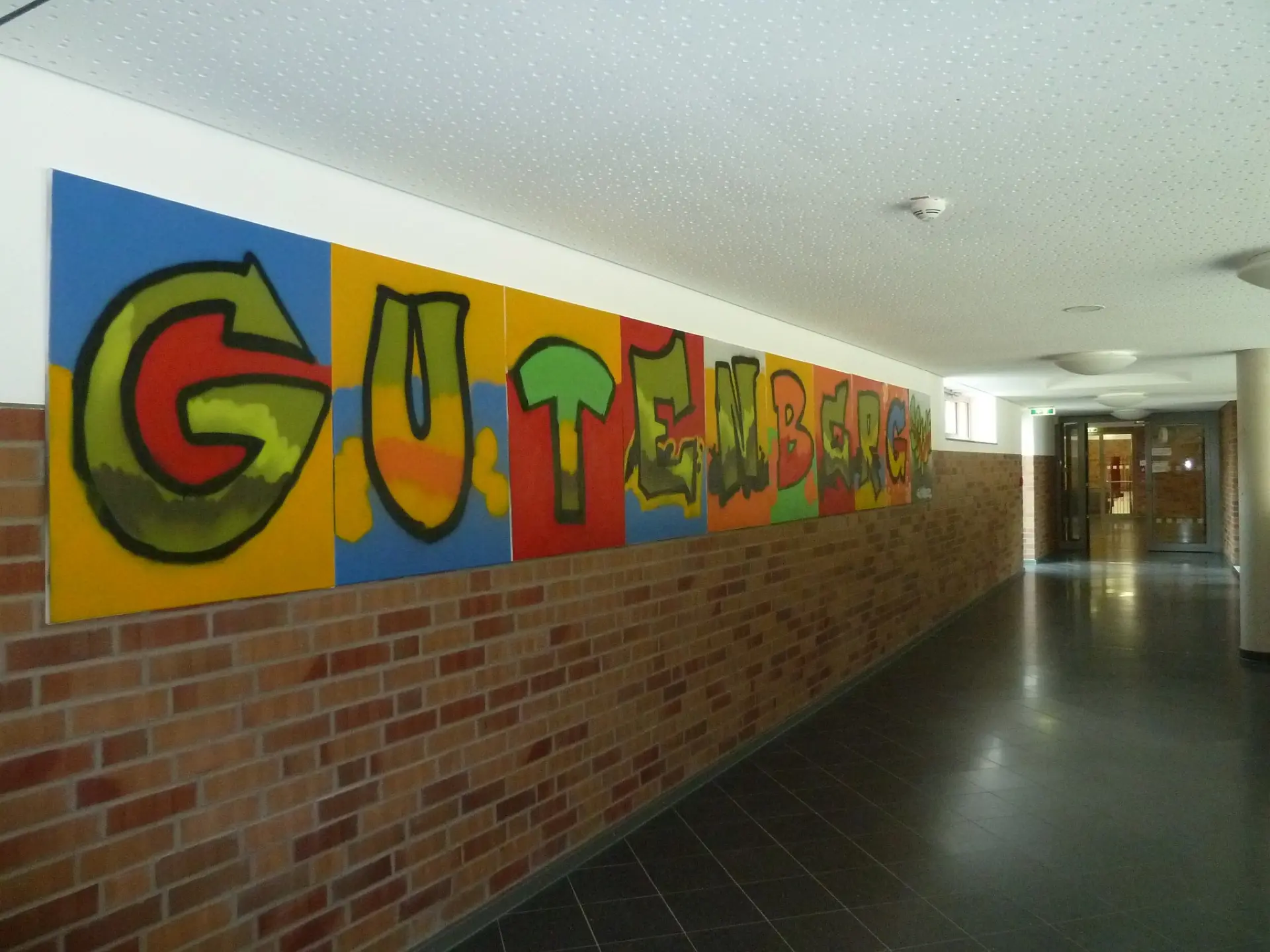 Ein langer Schulflur mit Backsteinwand, an der ein großformatiges, farbenfrohes Graffiti mit dem Schriftzug "GUTENBERG" angebracht ist