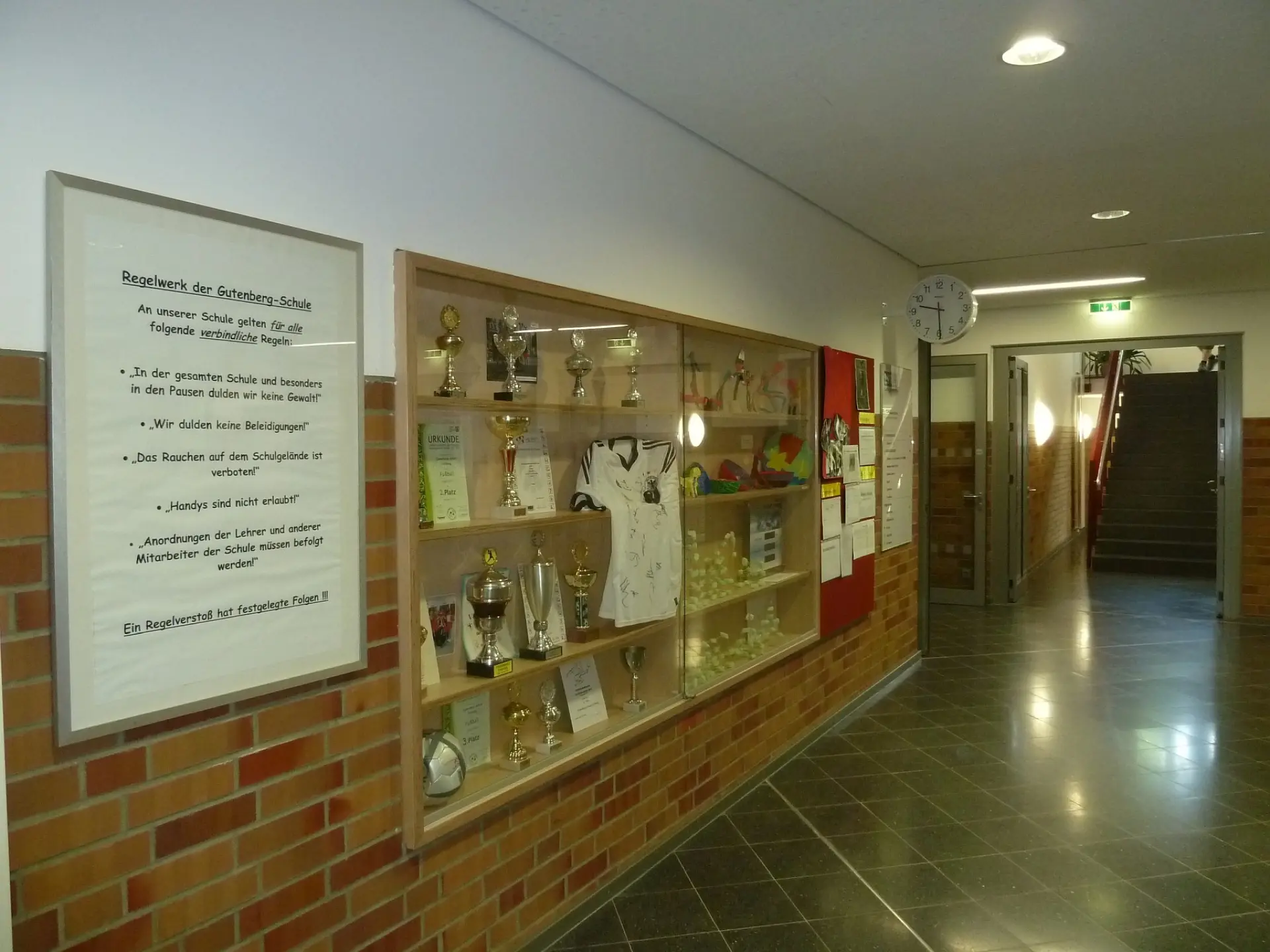 Schulflur mit einer Glasvitrine für Pokale und Auszeichnungen sowie einem gerahmten Aushang mit dem "Regelwerk der LVR-Gutenberg-Schule"