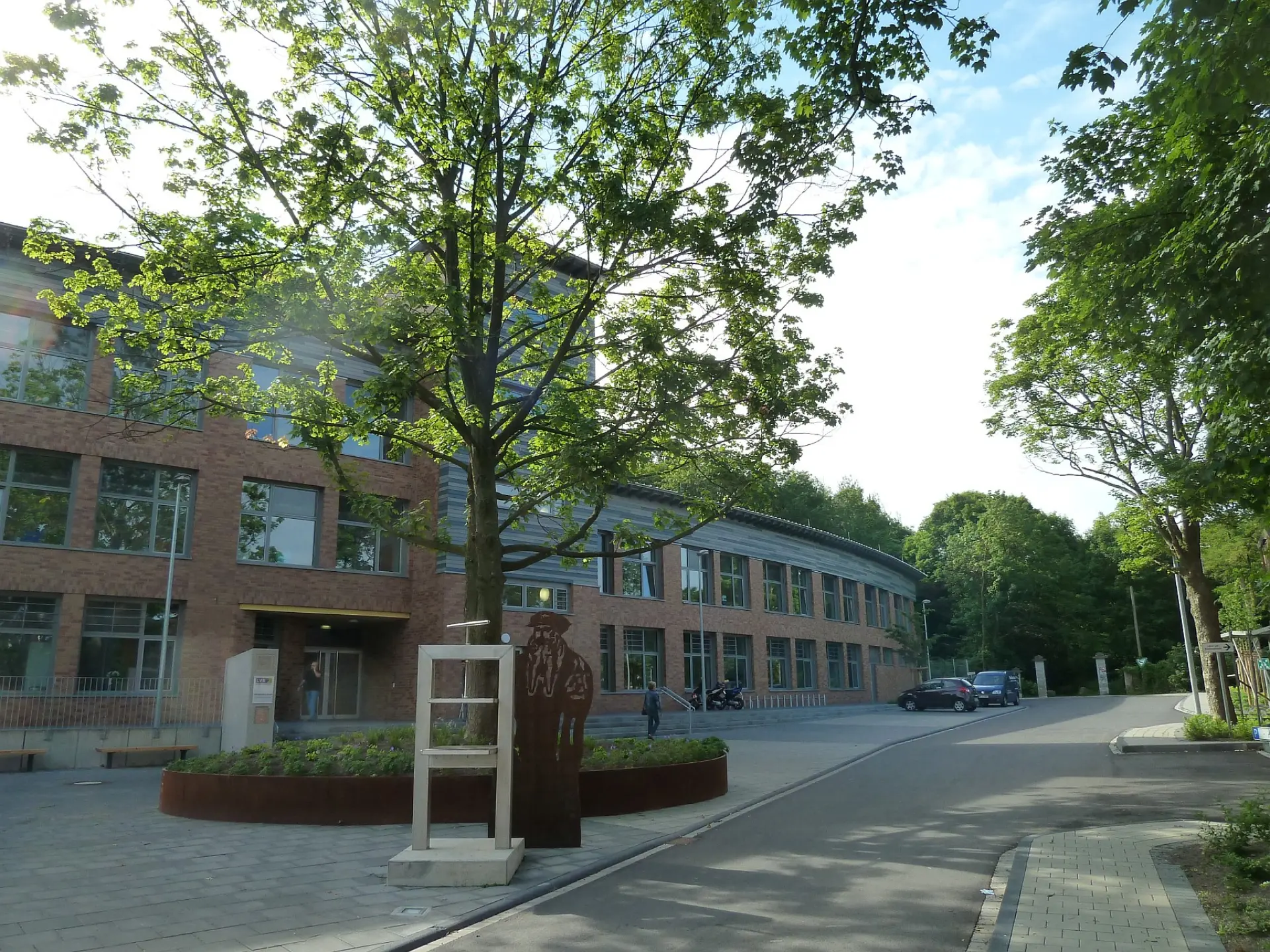 Haupteingang der LVR-Gutenberg-Schule mit einer modernen Backsteinfassade, einem großen Baum im Vorhof und einer Metall-Skulptur des Namensgebers Johannes Gutenberg