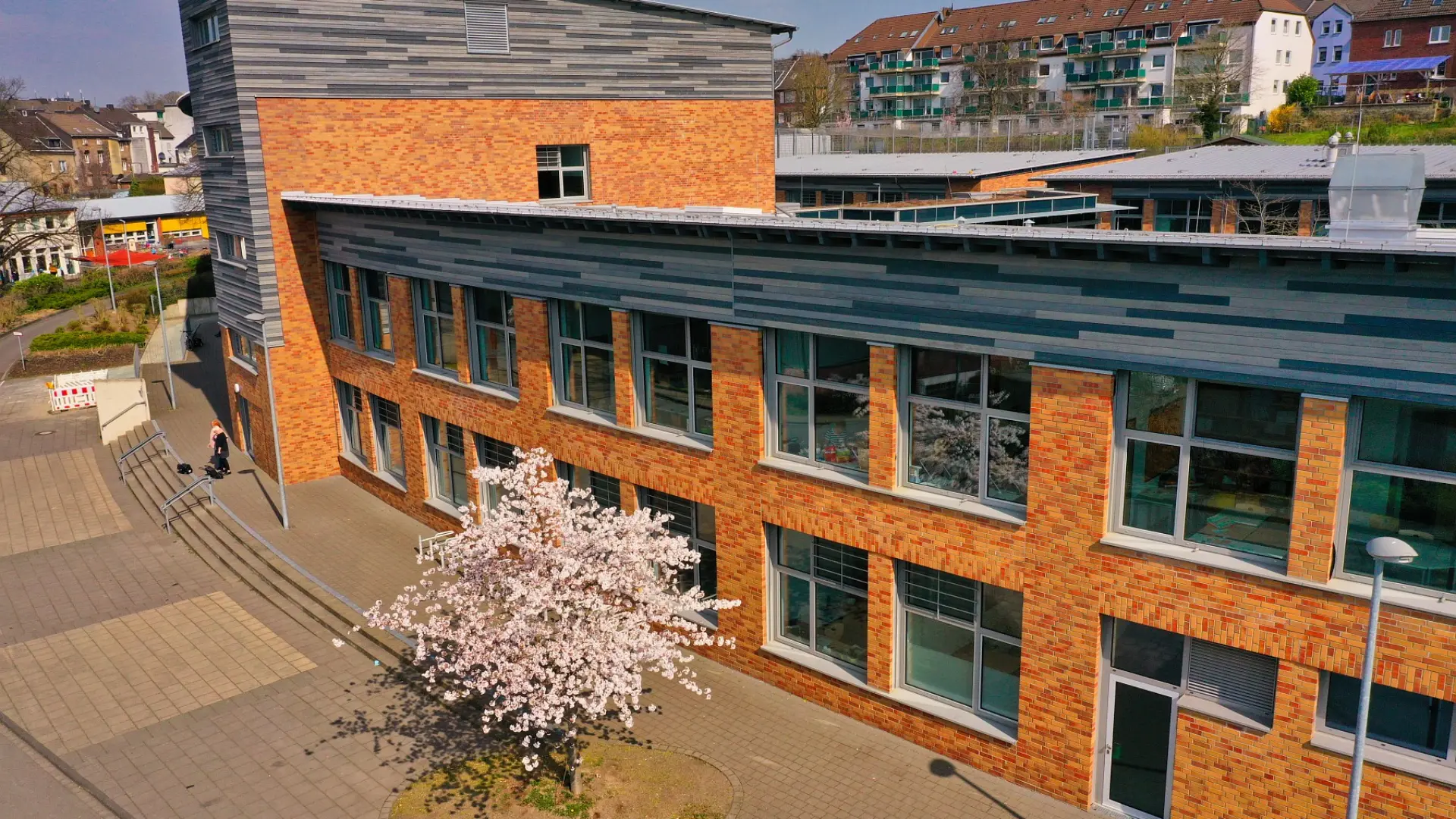 Luftaufnahme der LVR-Gutenberg-Schule mit Fokus auf die Backsteinfassade, ein markantes Treppenhaus-Element und einen blühenden Kirschbaum im Vordergrund