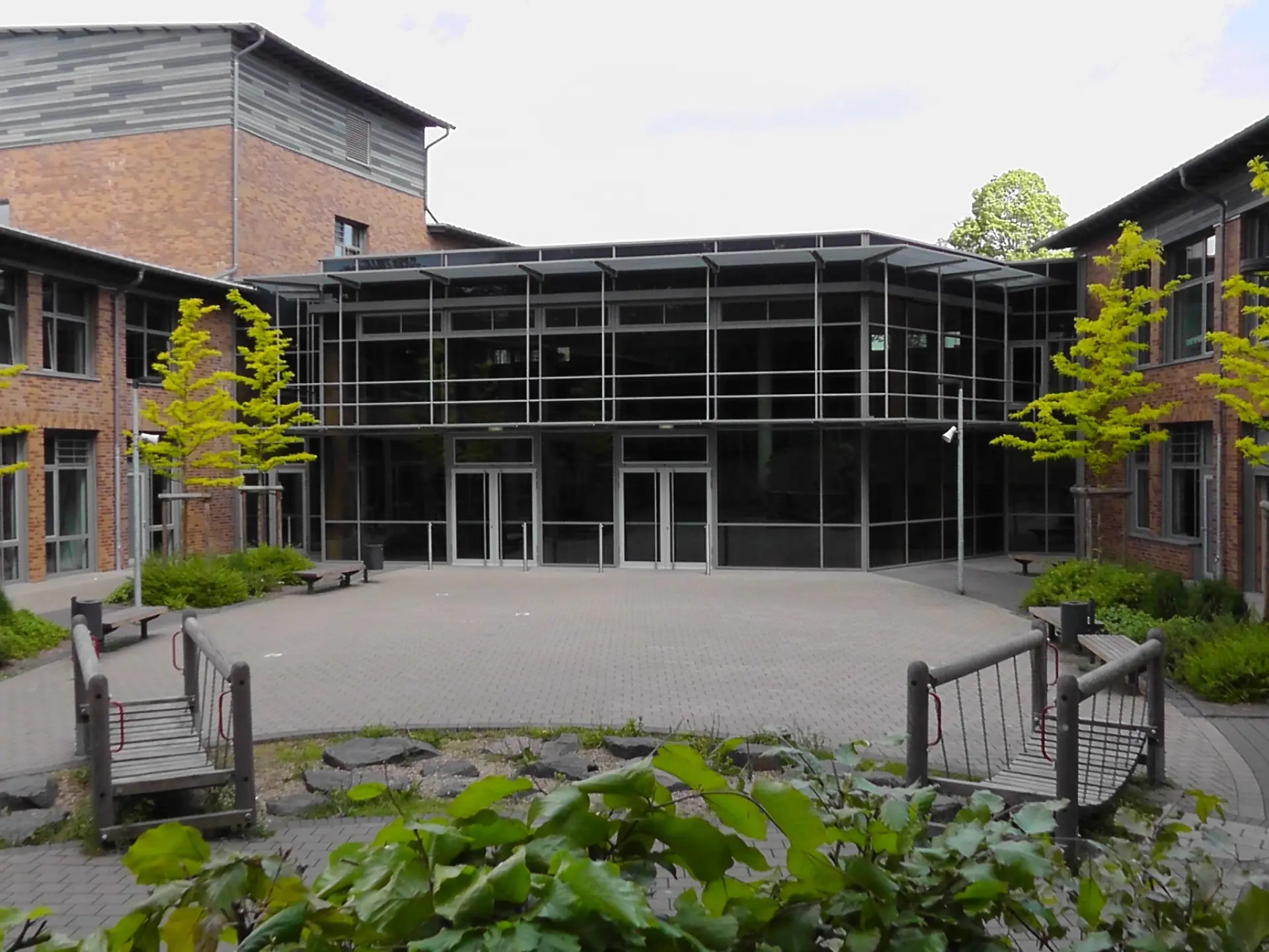 Gepflasterter Schulinnenhof mit zwei kleinen Holz-Hängebrücken für Kinder und einer modernen Glasfassade im Hintergrund