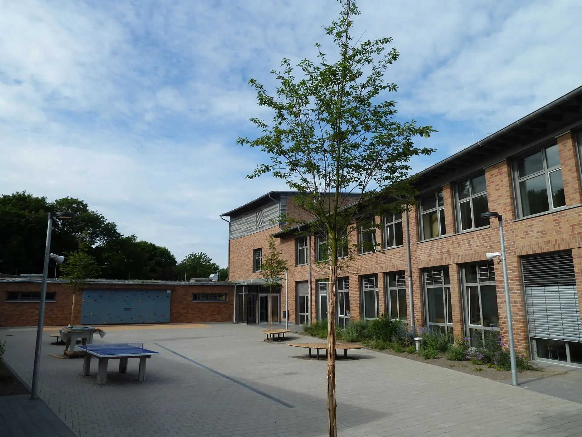 Schulinnenhof der LVR-Gutenberg-Schule mit Tischtennisplatten, Rundbänken unter jungen Bäumen und einer Kletterwand im Hintergrund