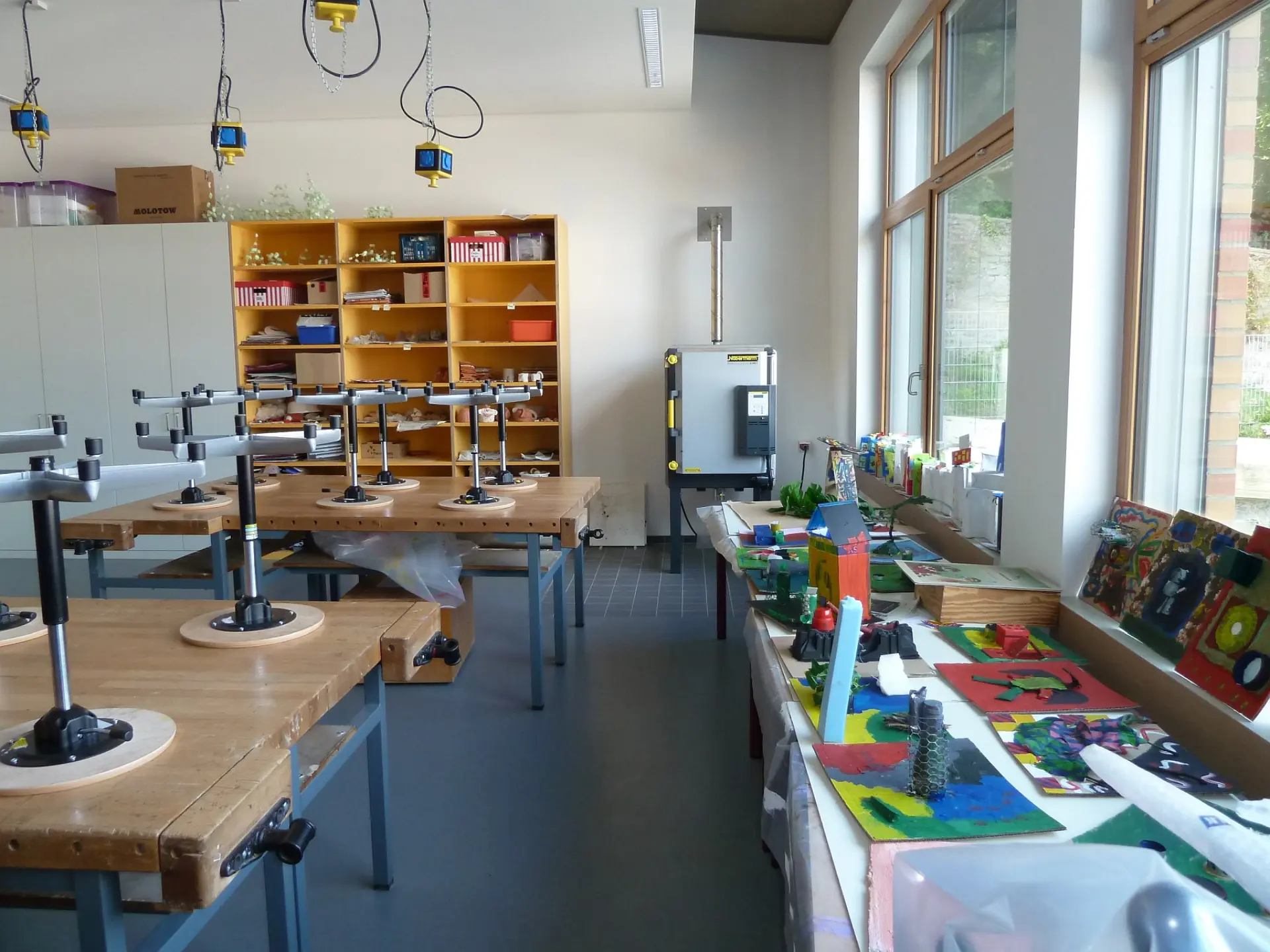 Lichtdurchfluteter Kunstraum mit großen Werktischen, Regalen voller Materialien und ausgestellten Arbeiten der Schüler*innen auf einer Fensterbank