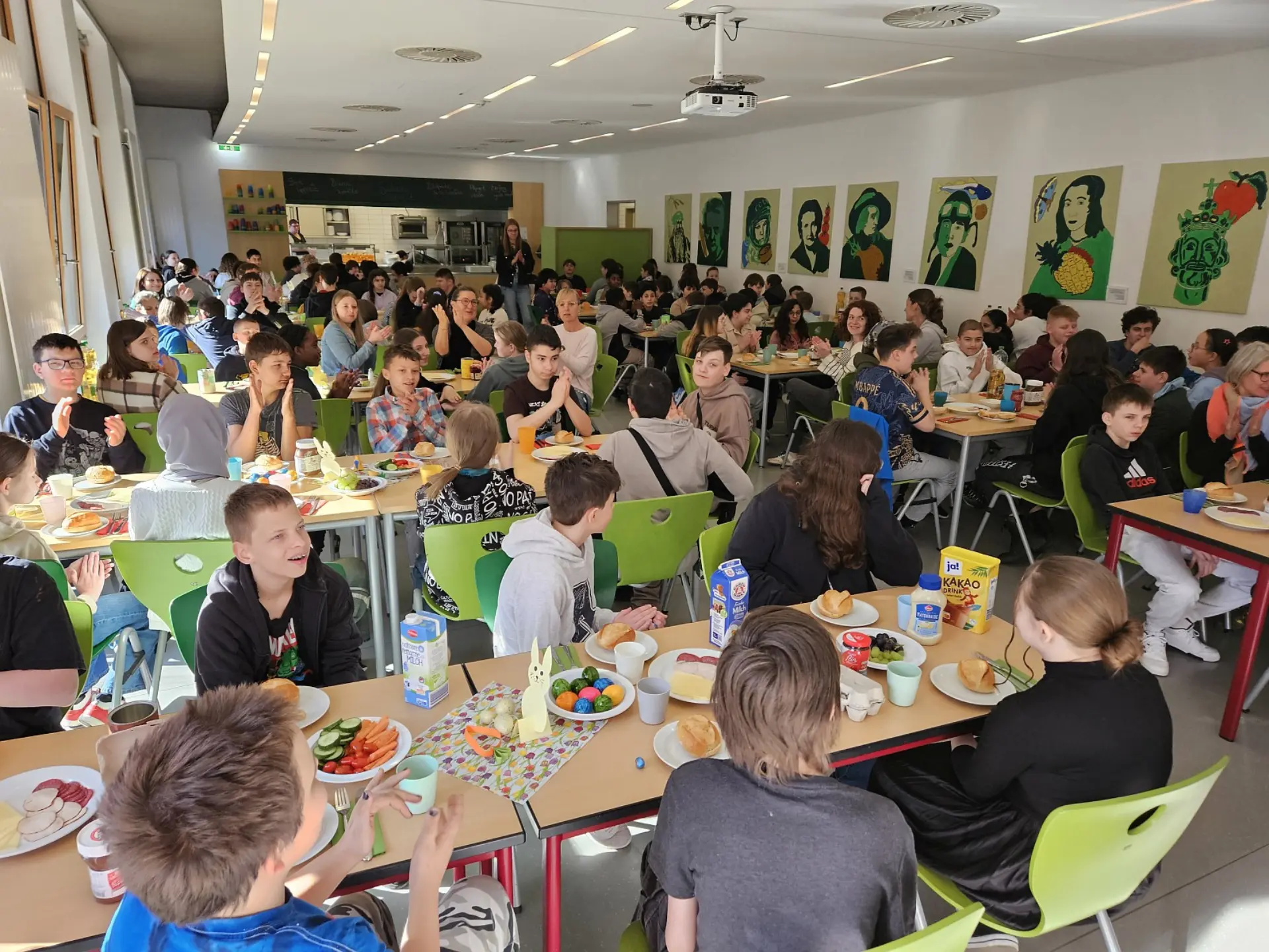 Die volle Schulmensa an einem sonnigen Tag. Alle Tische sind besetzt mit Kindern aus der Mittelstufe. Auf jedem Tisch gibt es Teller, Tassen, Besteck und Zutaten für ein feierliches Osterfrühstück.