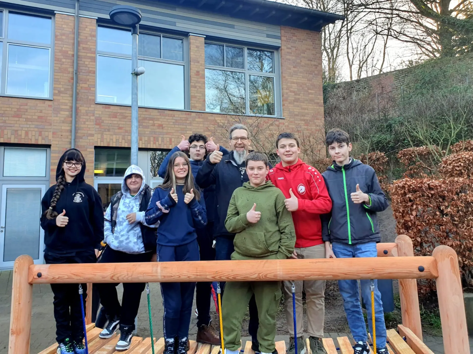 Eine Gruppe von sechs Schüler*innen und dem Schulleiter stehen auf einer neuwertigen Hängebrücke im Kletterbereich des Schulhofs. Sie alle blicken in die Kamera, lächeln und haben den Daumen hoch.