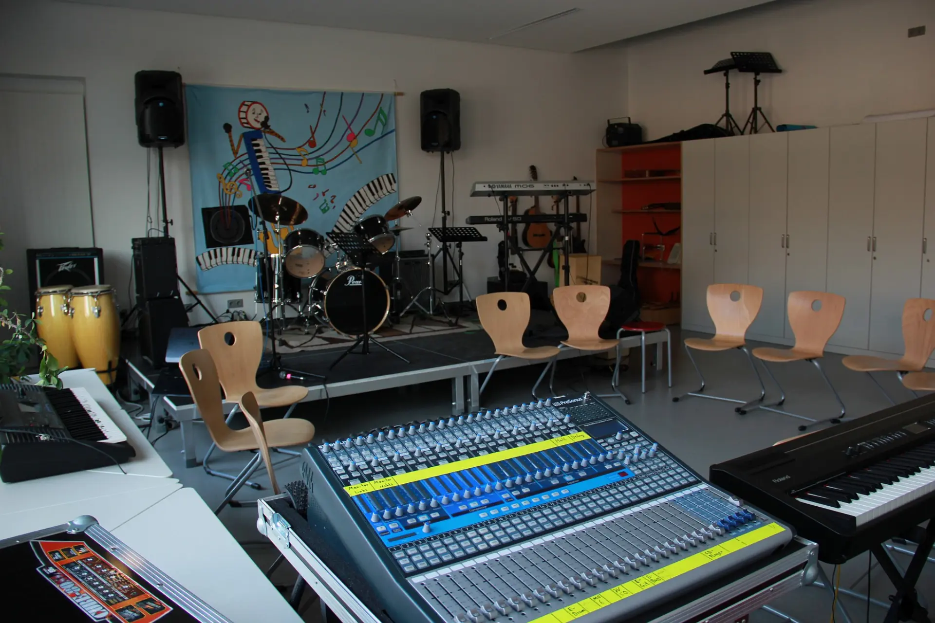 Moderner Proberaum der Schulband mit Schlagzeug auf einer Bühne, Mischpult im Vordergrund sowie Keyboards und Gitarren