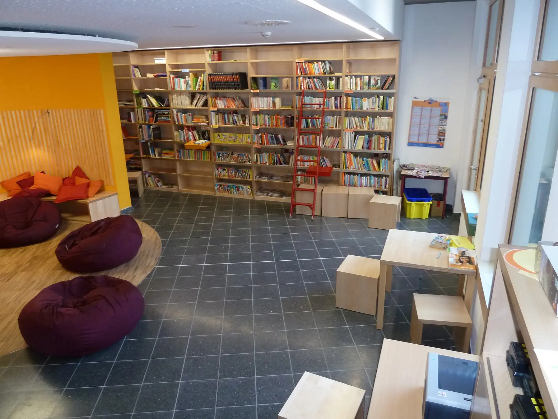 Blick in die helle Schulbibliothek: Eine raumhohe Holzwand ist komplett mit Büchern gefüllt, davor steht eine Bibliotheksleiter. Links lädt ein gemütlicher Bereich mit weichen Sitzsäcken und Kissen zum Lesen ein. Kleine Holztische und Würfelhocker bieten zusätzliche Arbeitsplätze.