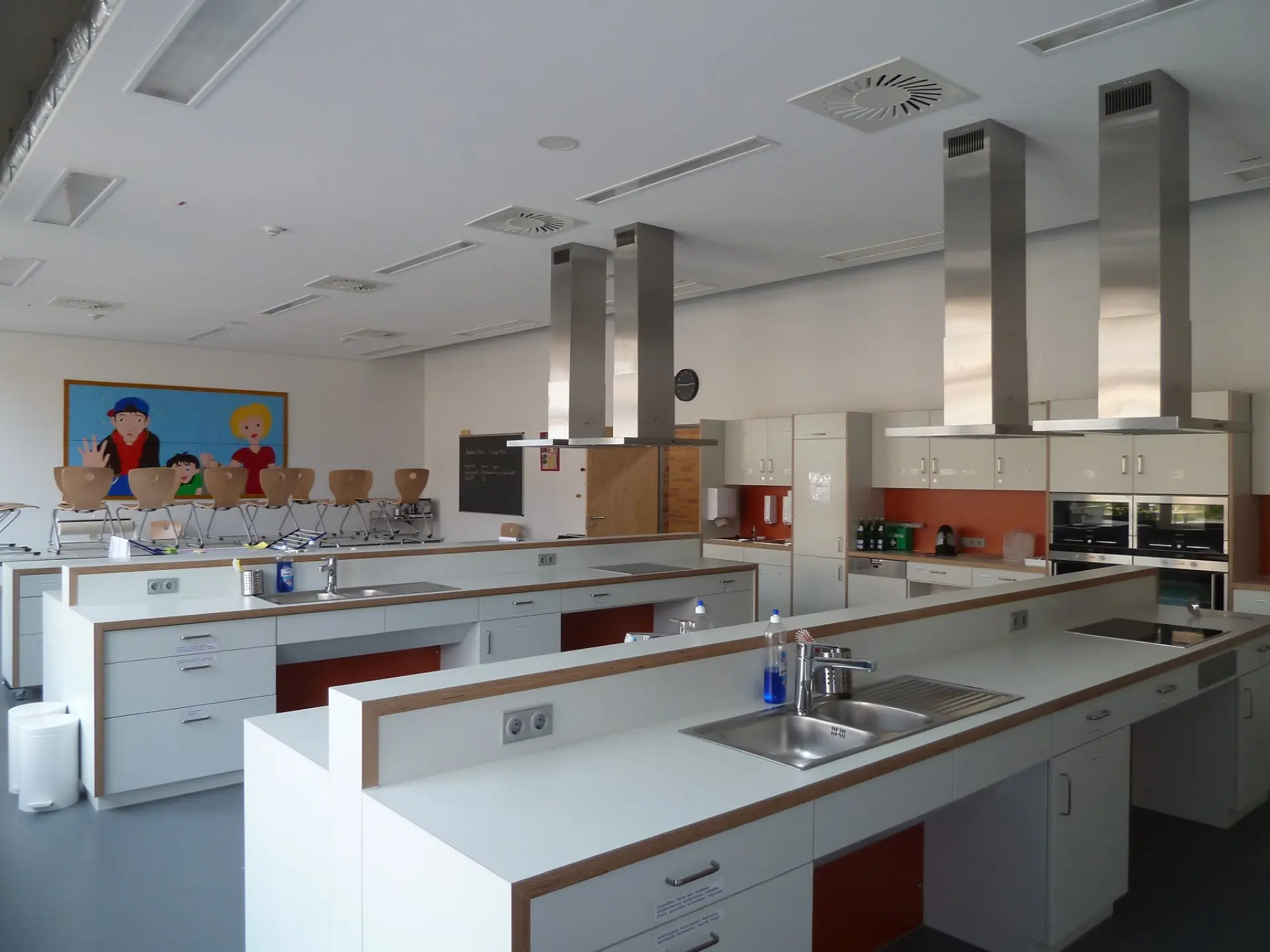 In der Schullehrküche stehen mehrere Kochinseln, ausgestattet mit Kochfeldern und Spülen. Über jeder Station hängt eine Dunstabzugshaube aus Edelstahl. Im Hintergrund sind weitere Küchenzeilen mit Backöfen sowie ein buntes Wandbild mit Comic-Figuren zu sehen.