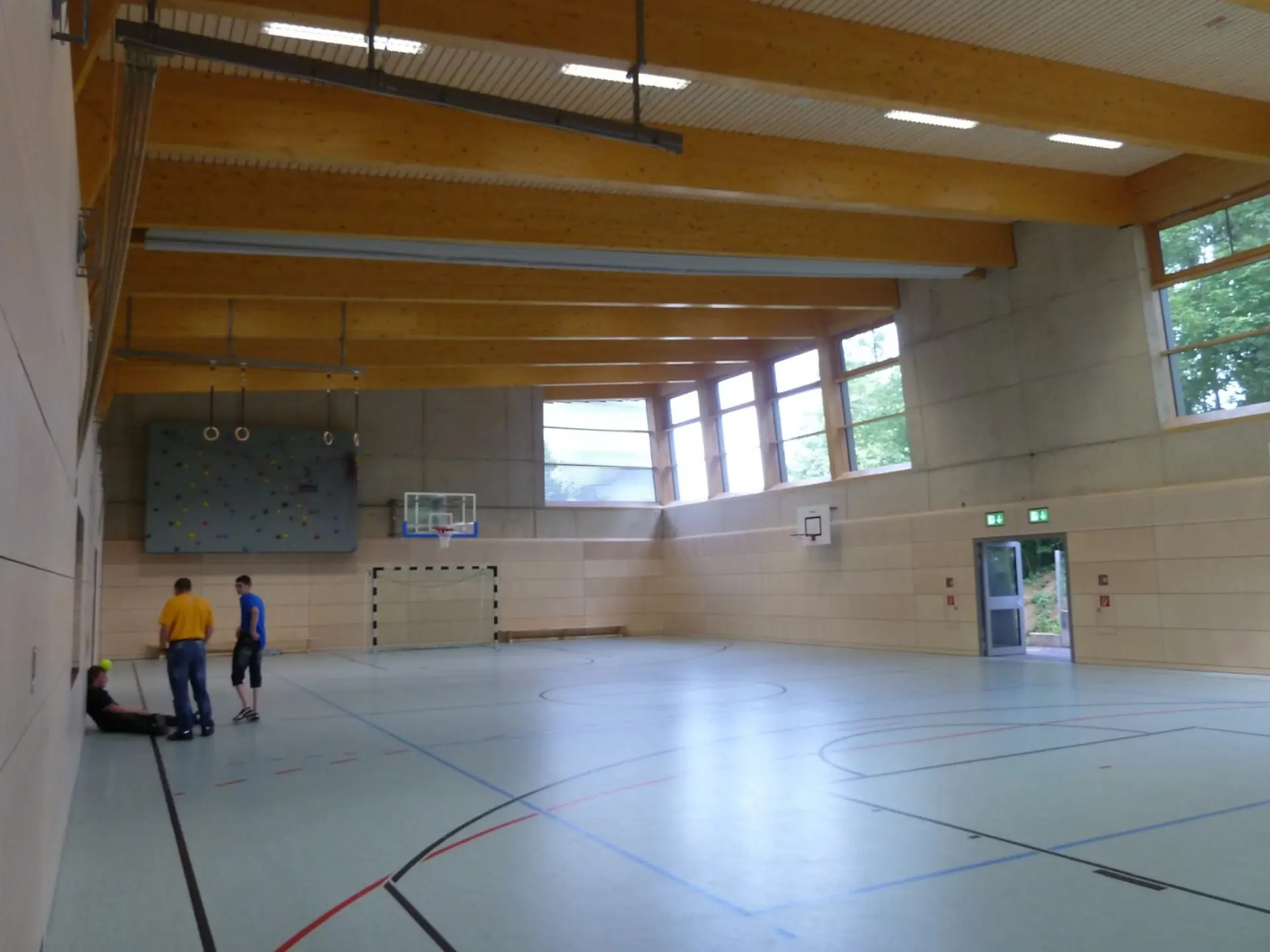 Große, moderne Sporthalle mit Holzbalkendecke, Basketballkörben, Fußballtor und einer Kletterwand an der Stirnseite