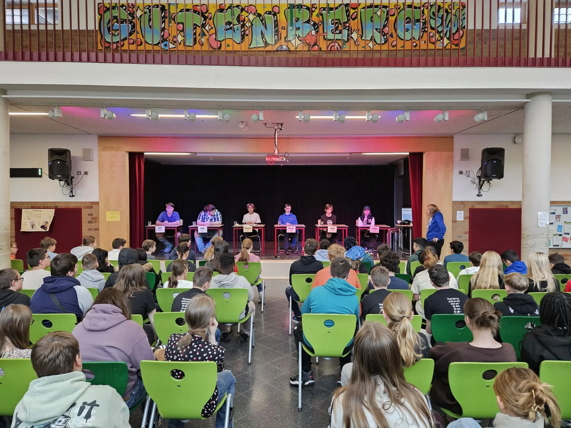Fünf Schüler*innen sitzen auf jeweils einem Einzeltisch auf der Bühne der Schulaula. Die Publikumsplätze vor ihnen sind komplett besetzt und alle hören ihnen zu.