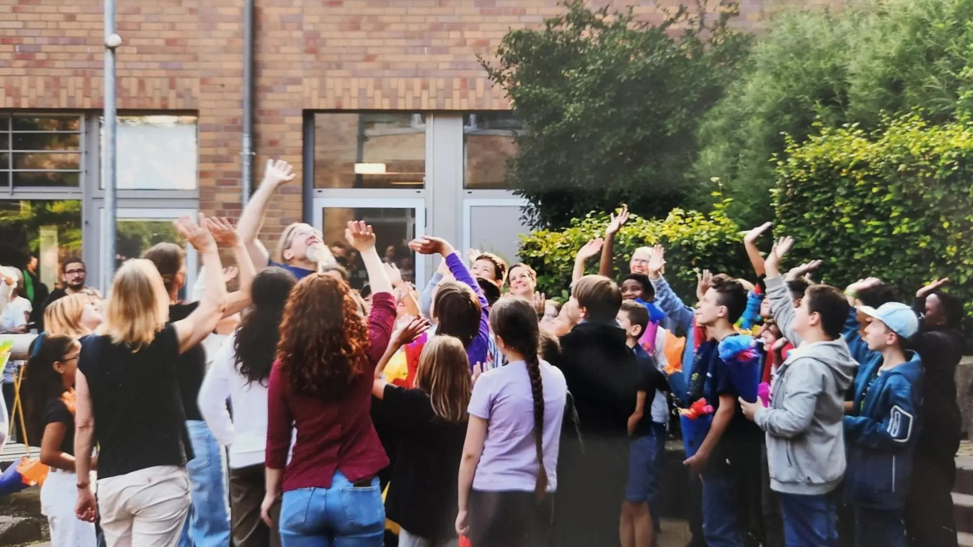 Eine große Gruppe von Schüler*innen und der Schulleiter stehen an einem Sommertag draußen vor dem Schulgebäude. Sie schauen nach oben zu einer über ihren Köpfen fliegenden Drohne (die nicht zu sehen ist), lächeln und winken in die Drohnenkamera.