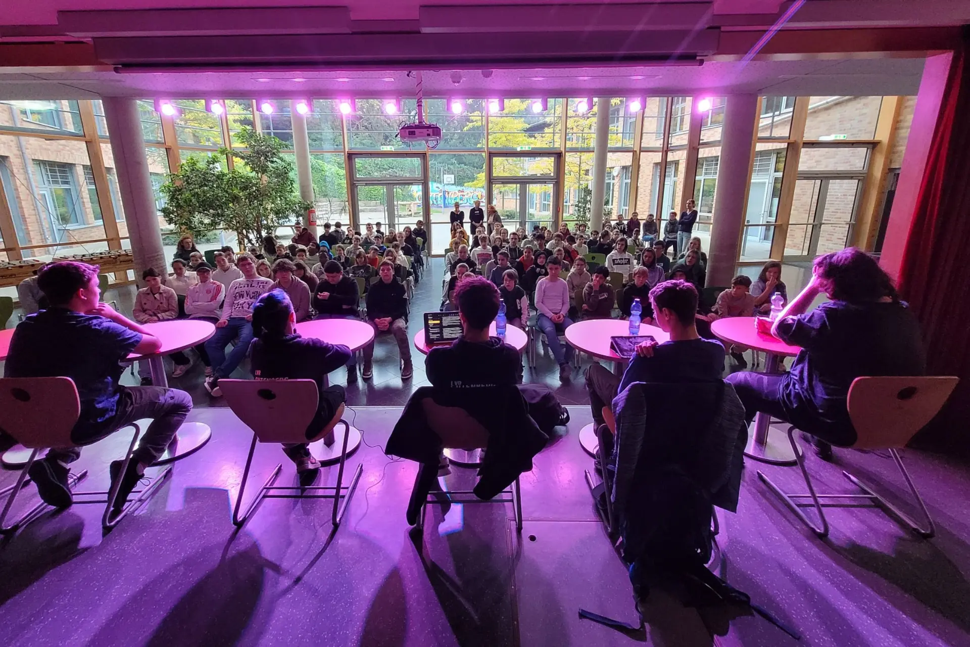 Auf der Bühne in der Schulaula sitzen fünf Schüler*innen. Sie werden von hinten aufgenommen, vor ihnen im vollen Saal sitzen viele weitere Schüler*innen und schauen sich die Diskussionsrunde an.
