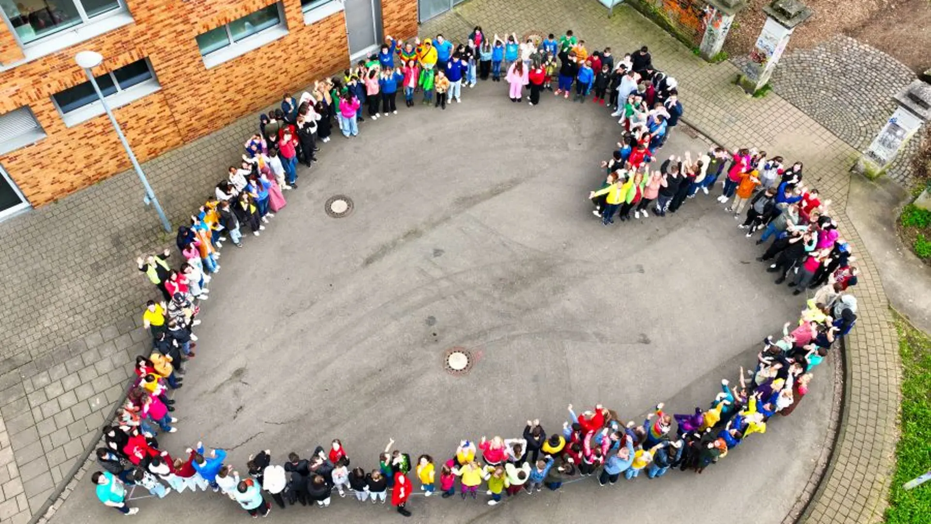 Eine Luftaufnahme zum Innenhof der Schule. Da hat sich eine große Gruppe aus Schüler*innen so positioniert, dass sie eine Herzform bilden. Manche schauen nach oben zur Kamera und winken.
