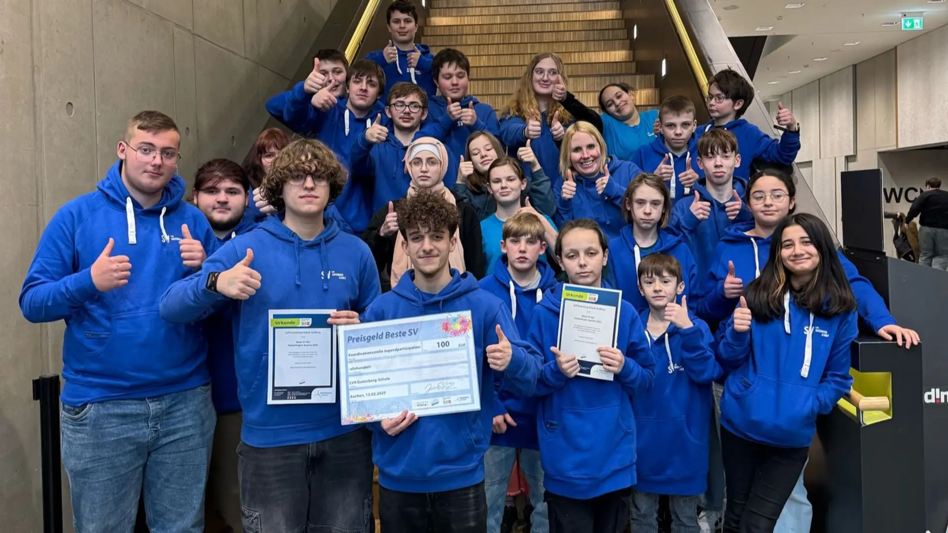 Eine Gruppe aus ca. 25 Schüler*innen steht an der Treppe in einem Schulgebäude. Alle schauen in die Kamera und lächeln, drei halten die Urkunden, die die Gruppe gewonnen hat, in der Hand. Alle tragen blaue Kapuzenpullis mit der Schrift LVR-Gutenberg-Schule darauf. 