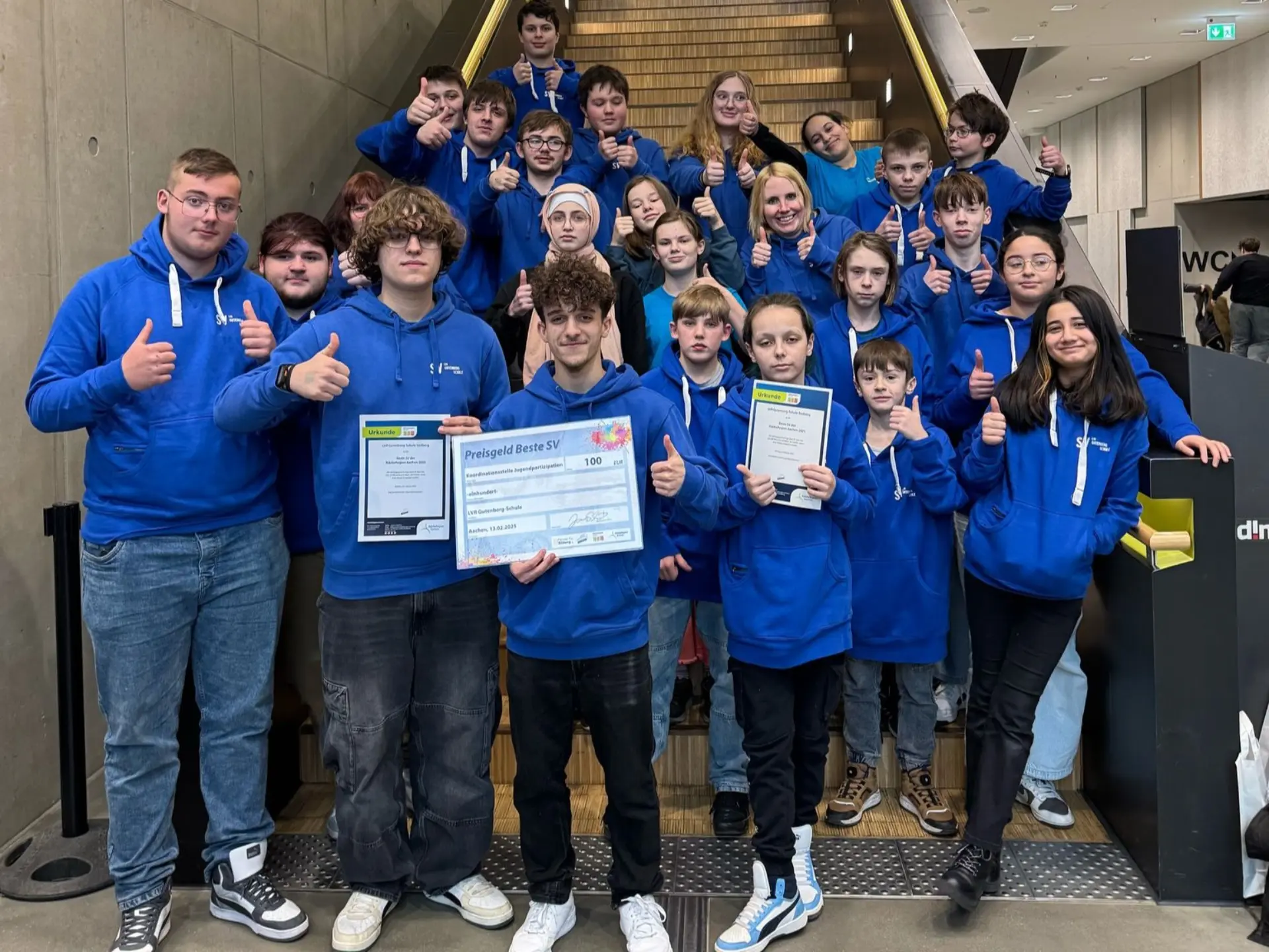 Eine Gruppe aus ca. 25 Schüler*innen steht an der Treppe in einem Schulgebäude. Alle schauen in die Kamera und lächeln, drei halten die Urkunden, die die Gruppe gewonnen hat, in der Hand. Alle tragen blaue Kapuzenpullis mit der Schrift LVR-Gutenberg-Schule darauf. 